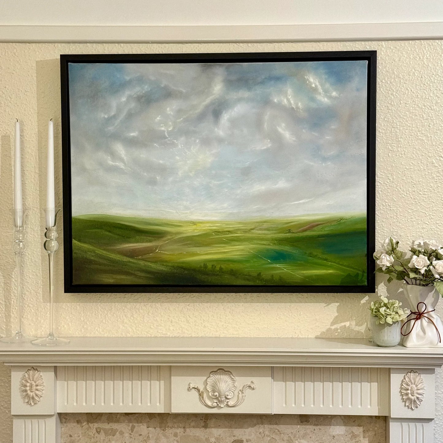 Framed landscape oil painting of green hills and dramatic sky displayed above a mantel