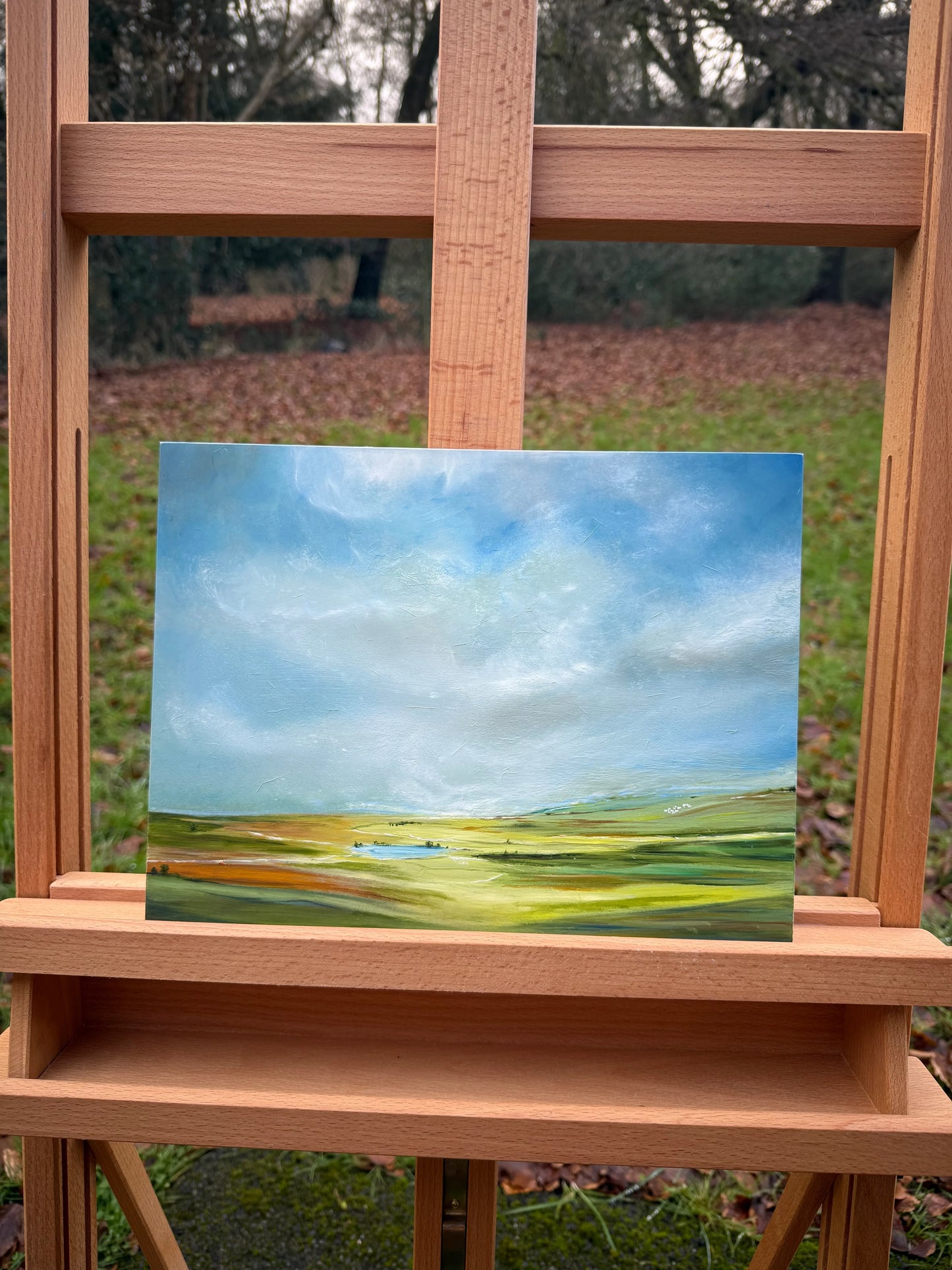 Romantic landscape oil painting with blue sky and green fields on a wooden easel outdoors.