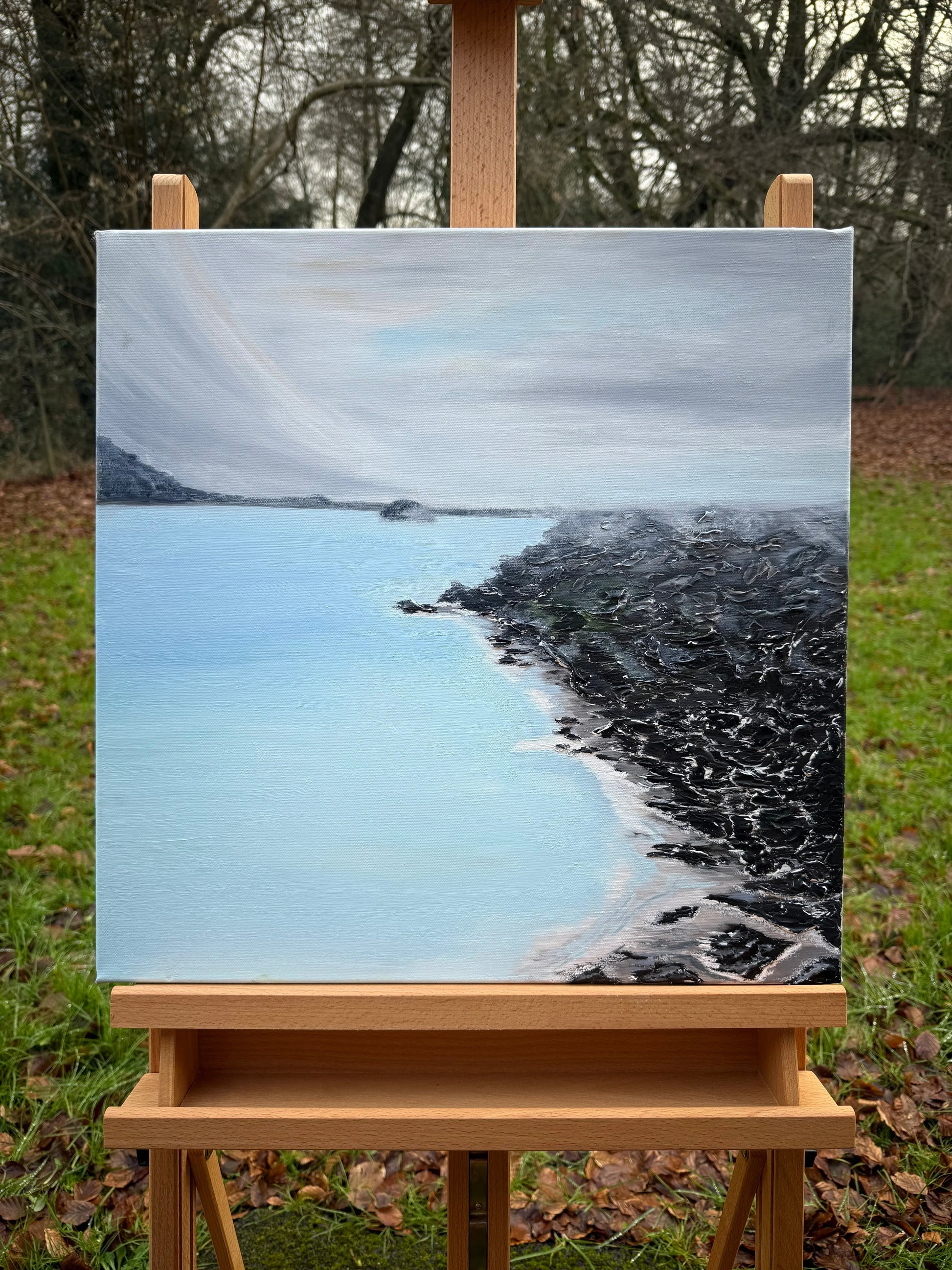 Romantic landscape oil painting of a tranquil blue lagoon with rocky shoreline on an easel.