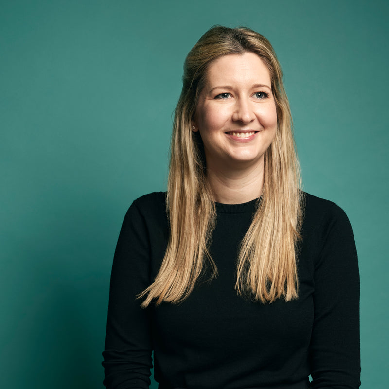 Woman with long blonde hair wearing a black jumper against a teal background
