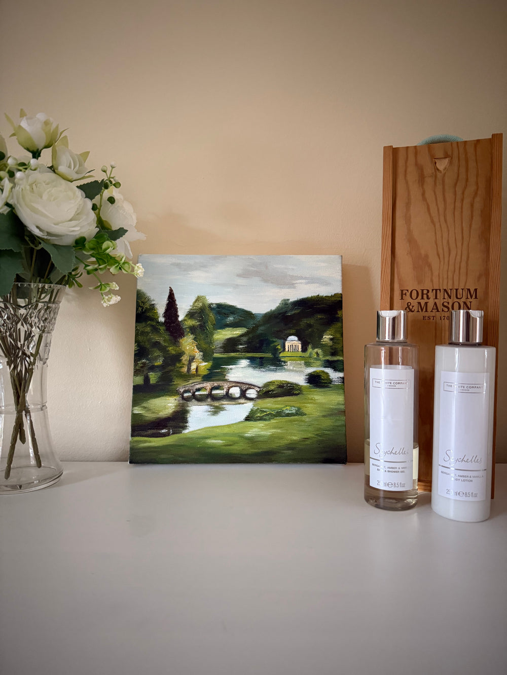 An oil painting of Stourhead (National Trust UK) on a table with flowers, fortnum and mason wine box and the white company toiletries