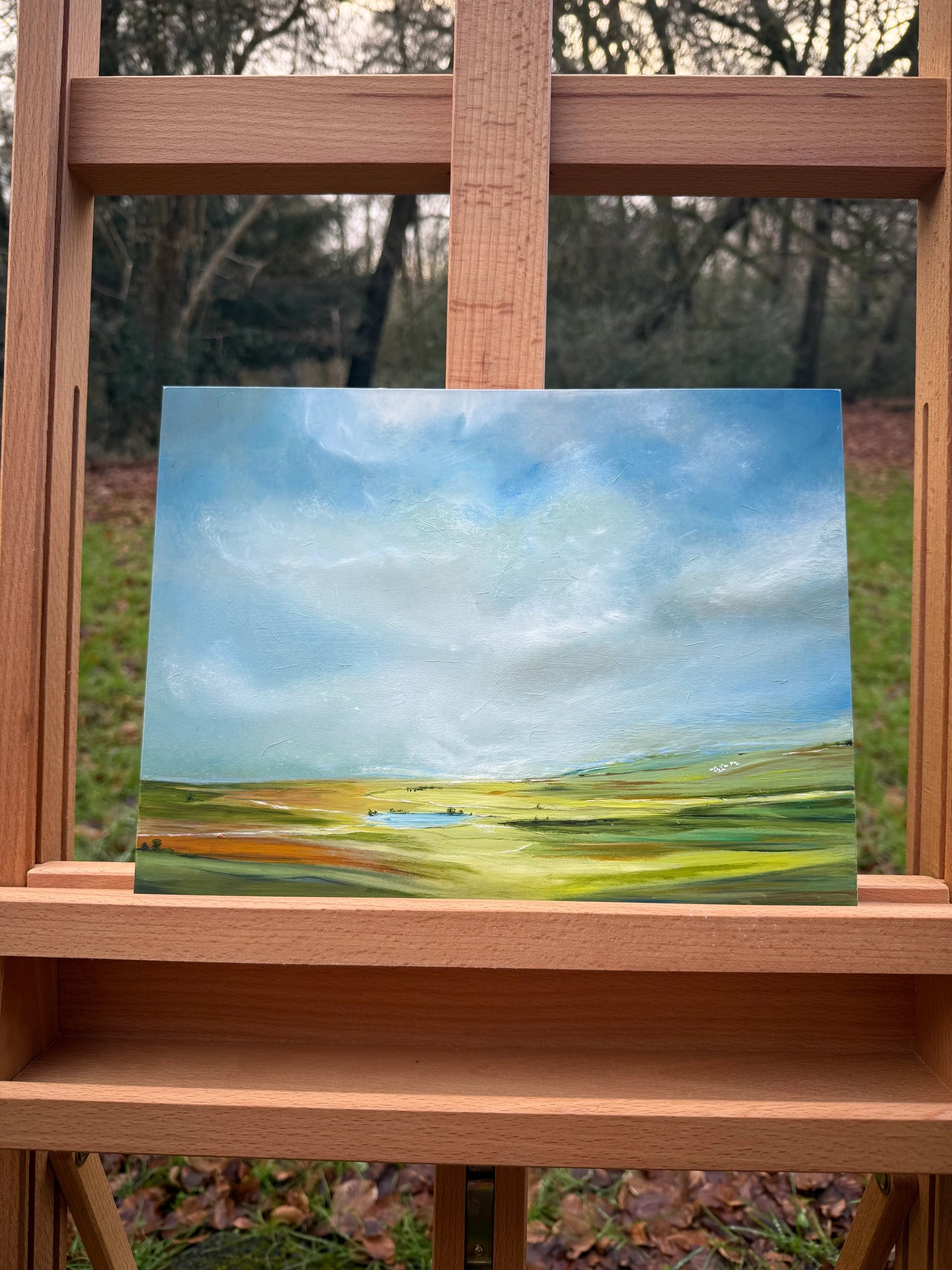 Oil painting of a peaceful landscape with grassy fields, blue sky, and small lake on easel outdoors.