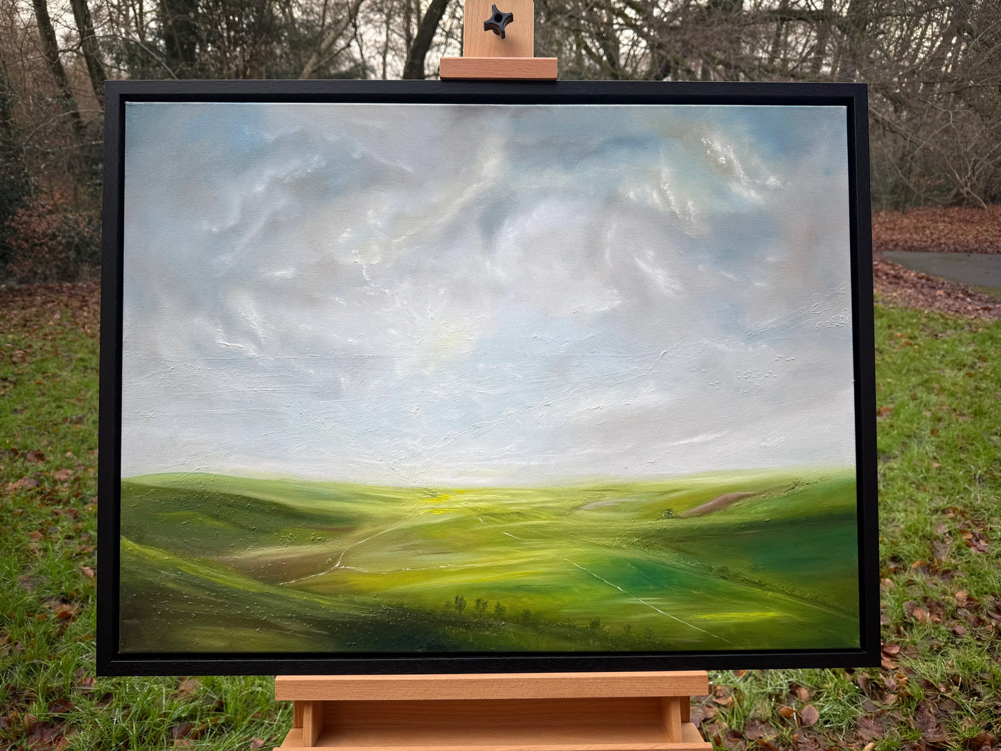 Framed oil painting of a serene green landscape under a bright, cloudy sky on an easel outdoors