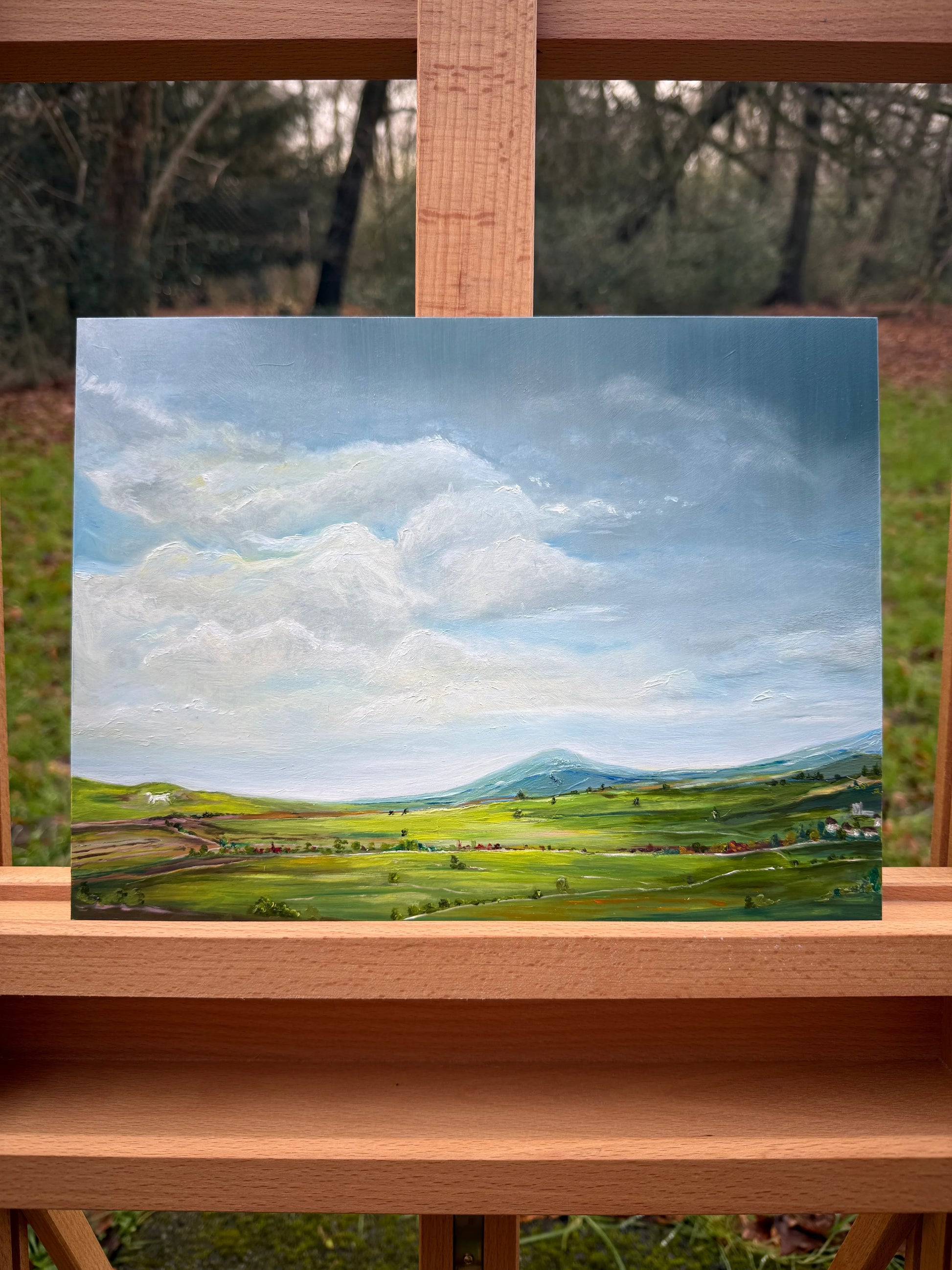 Romantic oil painting of a peaceful green landscape with rolling hills and sky on an easel.
